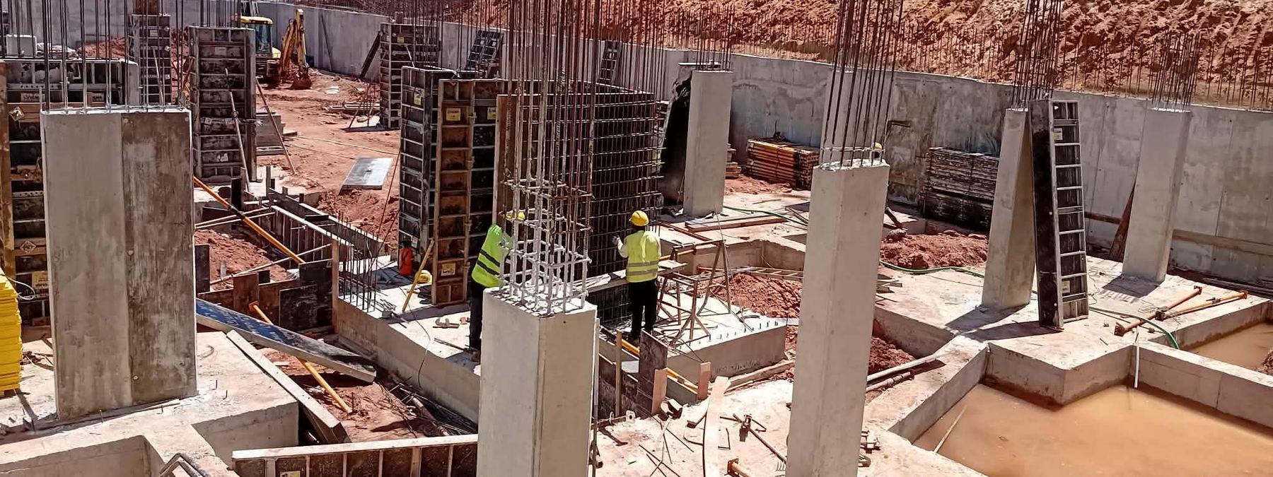 Obra com pilares em betão armado, armaduras verticais e muro de contenção, com equipa a montar cofragem